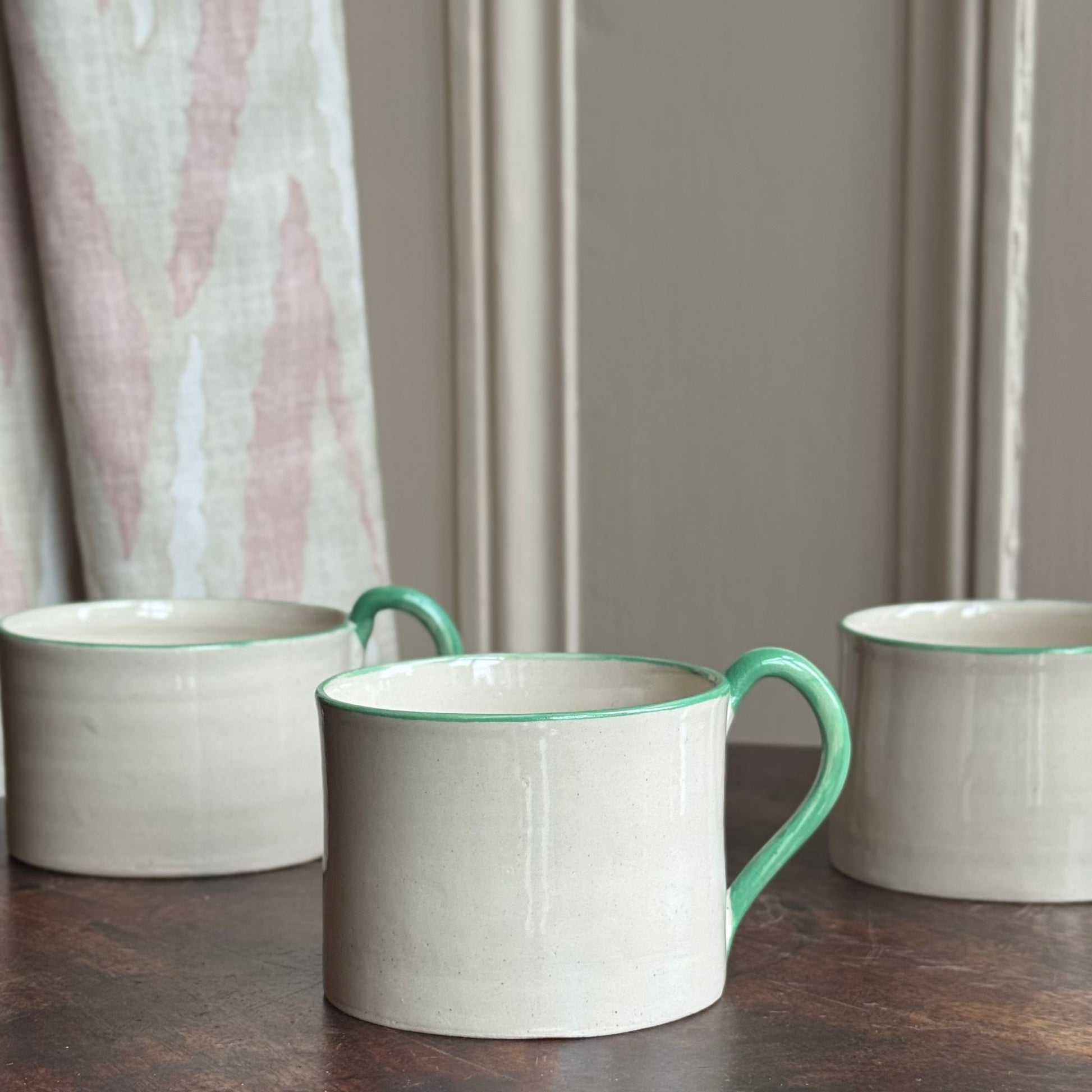 Three ceramic mugs with green rims on a wooden surface, with a neutral background.