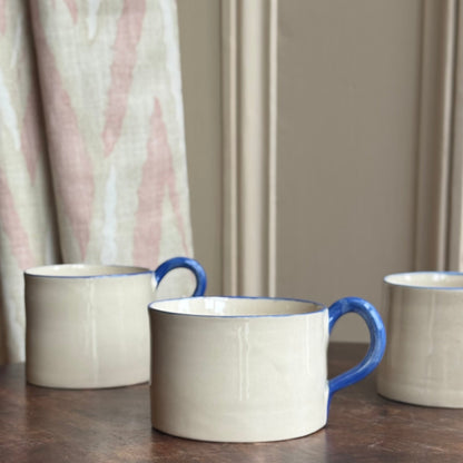Three handmade ceramic mugs with blue handles on a wooden table against a neutral wall.