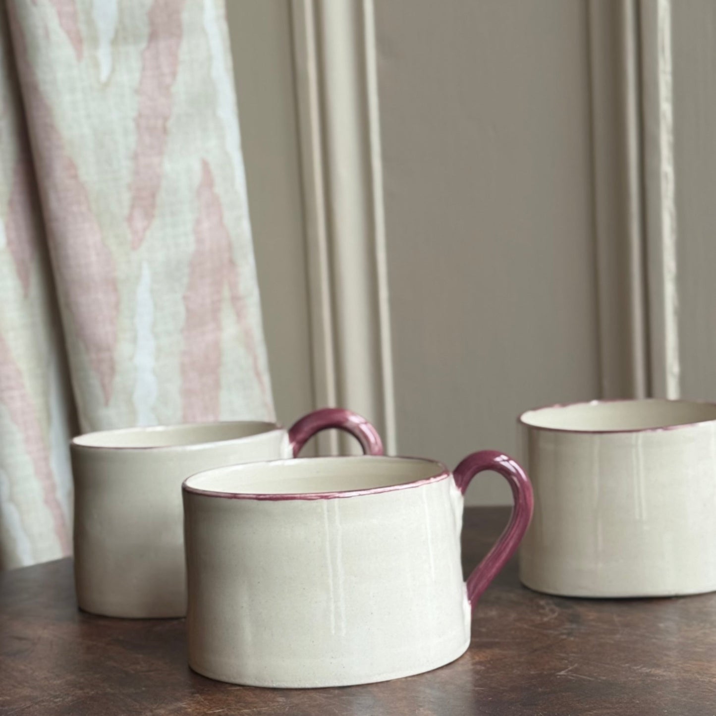 Three ceramic mugs with burgundy handles on a wooden table against a neutral curtain background
