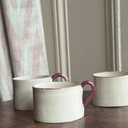 Three ceramic mugs with burgundy handles on a wooden table against a neutral curtain background