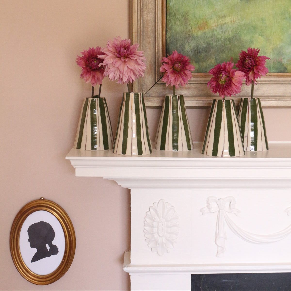 Five striped glass vases filled with pink flowers arranged on a white fireplace mantel against a peach-pink wall, with framed wall art in the background.