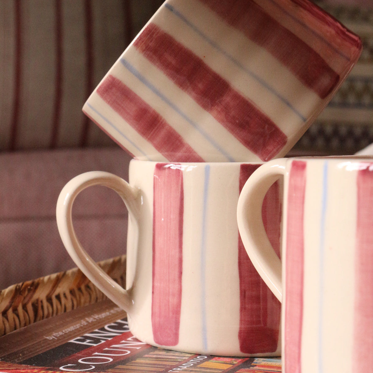 Three ceramic mugs with a cream base and burgundy and light blue stripes, styled on a tray with a book in a cozy indoor setting.